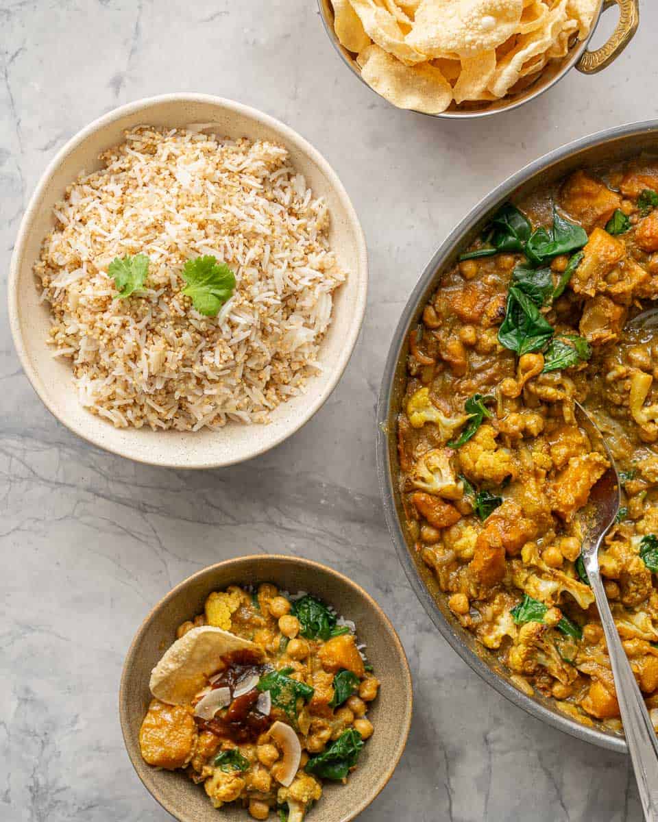 A bowl of quinoa rice, a skillet of curry, a bowl of poppadoms and a single serving of curry and toppings on a bench top. 